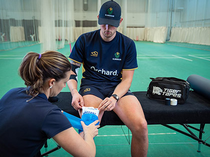 Georgina Ewer, Head Physiotherapist of Glamorgan Cricket, applying a hot/cold pack to a player's right knee, while he is sat on a therapy couch