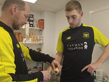 Gloucestershire cricket physiotherapist (left) using sports underwrap tape on a cricket player (right), both standing in a medical room with more tape in the background.
